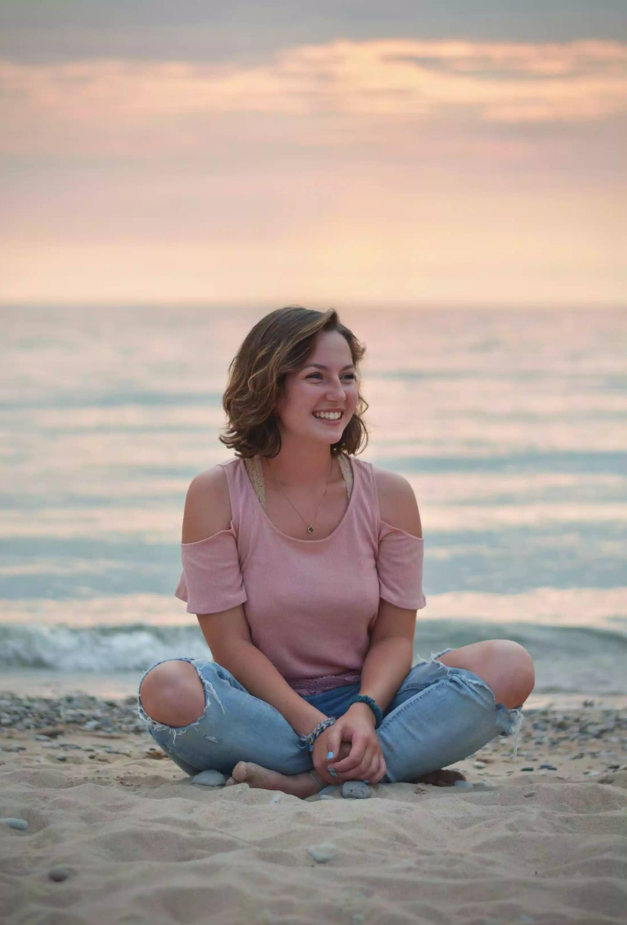 Girl sitting in the sand with a sunset behind her. She is wearing a pink t-shirt and blue jeans and smiling while facing to the left.